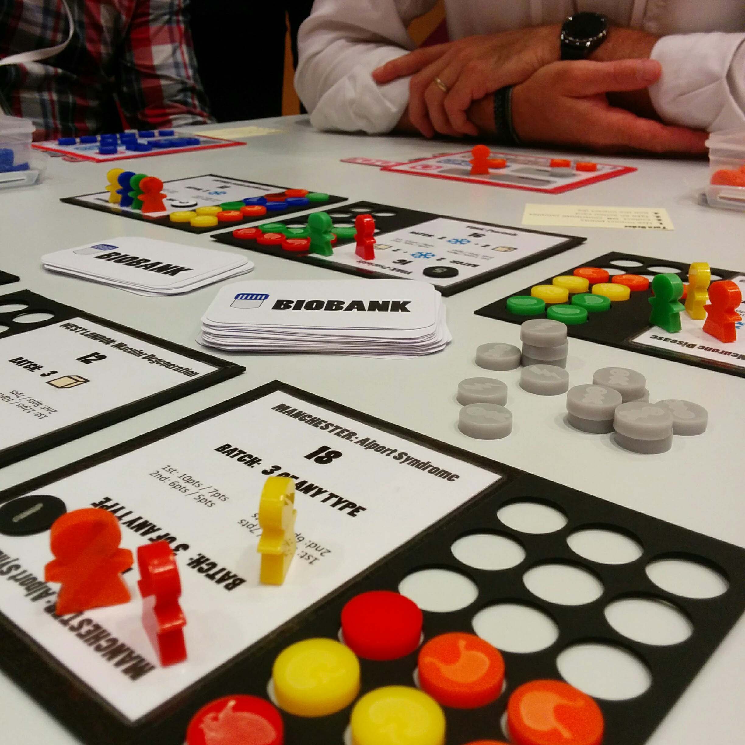 Biobank_EBW2016 A black player board for a board game that is half filled with with red and yellow circular pieces. There are also red and yellow meeples on the board. A person's folded arms can be seen in the background.