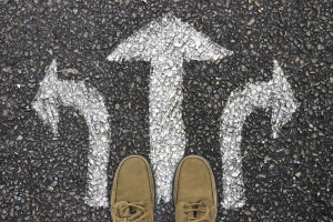 Photograph taken from above showing three arrows (one pointing left, one right and one straight ahead) drawn in white chalk on tarmac. A pair of feet in beige lace-up shoes is visible at the base of the three arrows.